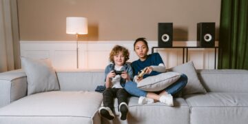a woman and a kid holding video game controllers while sitting on a sofa